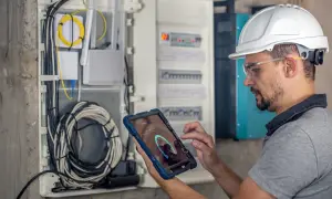 Elektryk w kasku i okularach ochronnych wykonuje przegląd instalacji elektrycznej, korzystając z tabletu z aplikacją diagnostyczną.