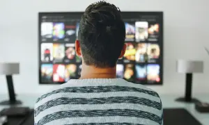 Mężczyzna w pasiastym swetrze ogląda na telewizorze menu aplikacji, gdzie widać okładki filmów i seriali.