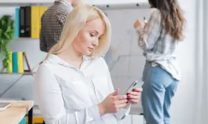 Kobieta z blond włosami korzysta z telefonu, być może używając wyszukiwania głosowego, podczas gdy koledzy pracują przy tablicy.