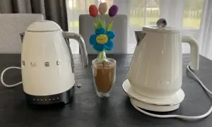 Białe czajniki elektryczne Smeg i DeLonghi, idealne do rankingu. Obok szklanka kawy i bukiet tulipanów.