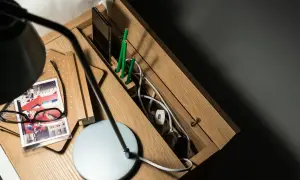 Drewniana przegródka na biurku z kablami, telefonem i zielonymi ołówkami. Widać też lampkę i okulary.