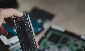 Dłoń trzyma moduł pamięci RAM, prawdopodobnie LPDDR5, z widocznymi układami scalonymi. W tle widać inne komponenty komputerowe.