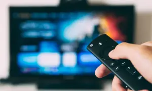 Dłoń trzymająca pilot do telewizora LG, ekran świeci w tle. Dowiedz się, jak wyłączyć lektora w telewizorze LG.