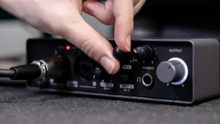 Dłoń obraca pokrętło MIX na panelu miksera audio. Widoczne wejście aux i inne złącza.