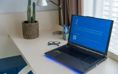 Laptop z niebieskim ekranem błędu formatu Windows 10 na biurku obok okularów i kaktusa.