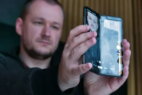 Mężczyzna trzymający składany smartfon, na ekranie którego widać jego twarz.