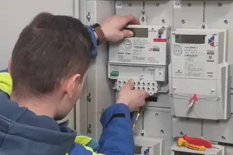 Technik w kamizelce wykonuje wymianę licznika Tauron. Widać nowe liczniki i narzędzia.