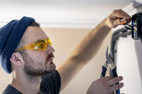 Elektryk w okularach ochronnych i czapce z wąsem ostrożnie podłącza przewody, zastanawiając się, z której strony faza w gniazdku.