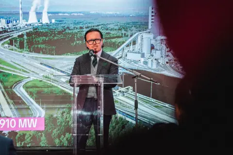 Mężczyzna przemawia na tle wizualizacji elektrowni i autostrady. Na ekranie widnieje napis "910 MW", symbolizujący zwiększenie mocy przyłączeniowej Tauron.