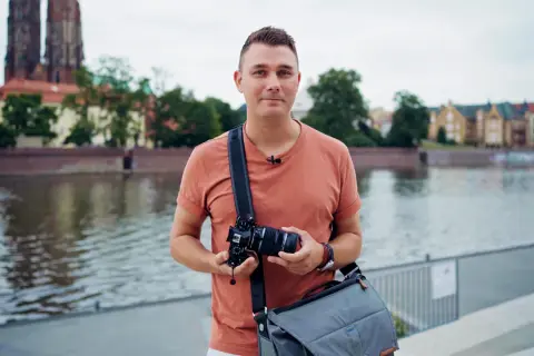 Mężczyzna w pomarańczowej koszulce trzyma aparat fotograficzny na tle rzeki i zabytkowych budynków.