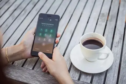 Zablokowany iPhone z ekranem wprowadzania kodu PIN, obok filiżanka kawy na drewnianym stole.