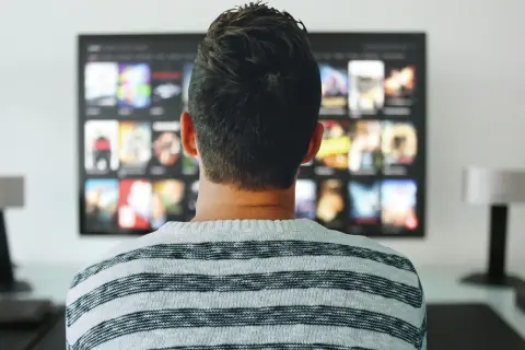 Mężczyzna w pasiastym swetrze ogląda na telewizorze menu aplikacji, gdzie widać okładki filmów i seriali.