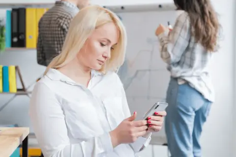 Kobieta z blond włosami korzysta z telefonu, być może używając wyszukiwania głosowego, podczas gdy koledzy pracują przy tablicy.