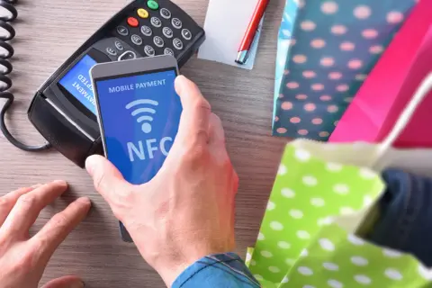 Płatność zbliżeniowa telefonem z funkcją NFC. Transakcja w toku, obok kolorowe torby na zakupy.