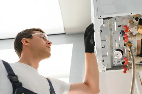 Instalator w okularach ochronnych sprawdza podłączenia kotła. Widać elementy takie jak anoda tytanowa i zawory.