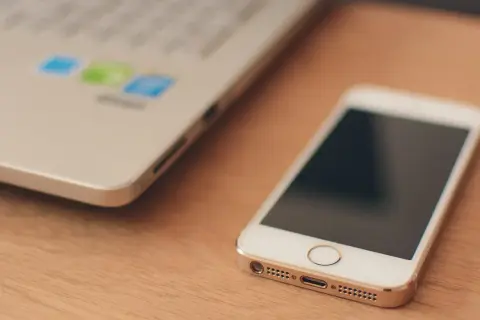 Biały smartfon leży obok laptopa. W tle widać logo z kolorowymi ikonami, sugerującymi uruchomione aplikacje w tle.