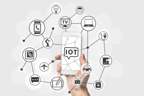 Dłoń trzyma smartfon z napisem "IOT co to". Wokół widnieją ikony urządzeń połączonych siecią: żarówka, TV, laptop, ekspres do kawy, pralka, samochód, aparat, samolot, robot, smartfon, router i chmura danych.