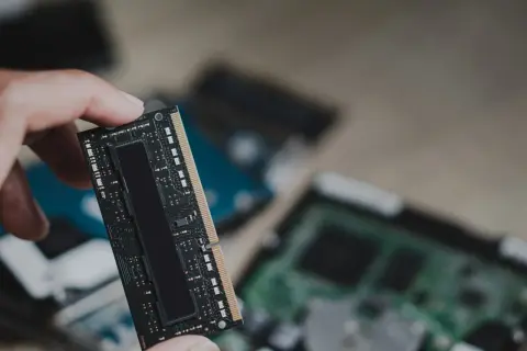 Dłoń trzyma moduł pamięci RAM, prawdopodobnie LPDDR5, z widocznymi układami scalonymi. W tle widać inne komponenty komputerowe.