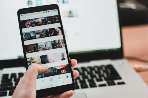Dłoń trzyma smartfon z widokiem na ekran z siatką zdjęć. Obok leży laptop. Czy to połączenie przez miracast?
