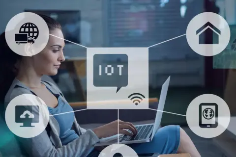Kobieta pracuje przy laptopie, otoczona ikonami symbolizującymi internet rzeczy (IoT): dom, chmura, ciężarówka, smartfon.