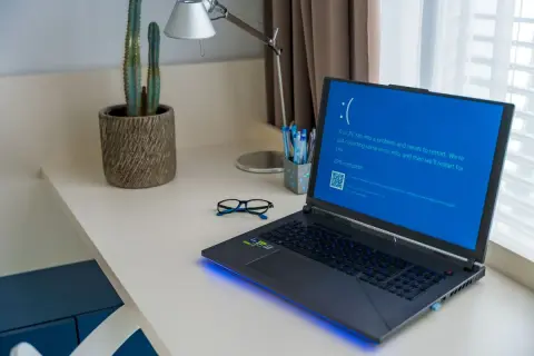 Laptop z niebieskim ekranem błędu formatu Windows 10 na biurku obok okularów i kaktusa.