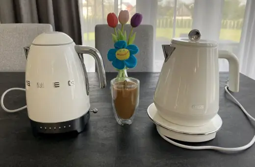 Białe czajniki elektryczne Smeg i DeLonghi, idealne do rankingu. Obok szklanka kawy i bukiet tulipanów.