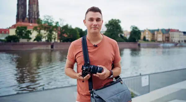 Mężczyzna w pomarańczowej koszulce trzyma aparat fotograficzny na tle rzeki i zabytkowych budynków.