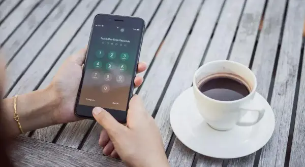 Zablokowany iPhone z ekranem wprowadzania kodu PIN, obok filiżanka kawy na drewnianym stole.