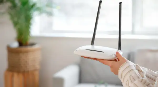 Dłoń trzyma biały router z dwiema antenkami. Może to być pierwszy krok, jak zresetować router, gdy internet przestaje działać.