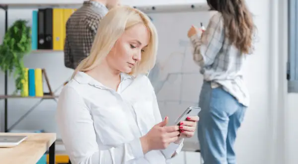 Kobieta z blond włosami korzysta z telefonu, być może używając wyszukiwania głosowego, podczas gdy koledzy pracują przy tablicy.