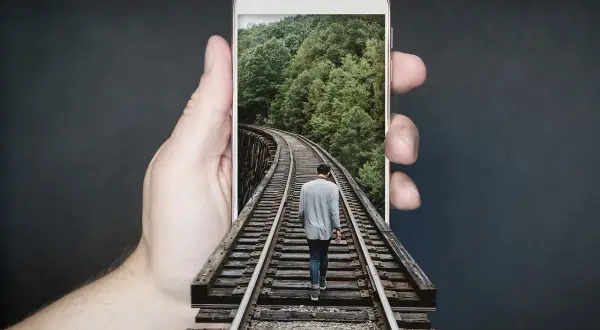 Dłoń trzyma telefon z ekranem **amoled**, na którym widać mężczyznę idącego po torach kolejowych przez las.