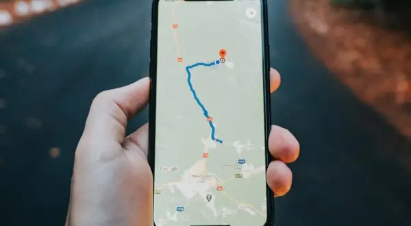 Dłoń trzyma iPhone'a z mapą Google, pokazującą trasę i lokalizację.