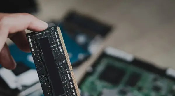 Dłoń trzyma moduł pamięci RAM, prawdopodobnie LPDDR5, z widocznymi układami scalonymi. W tle widać inne komponenty komputerowe.