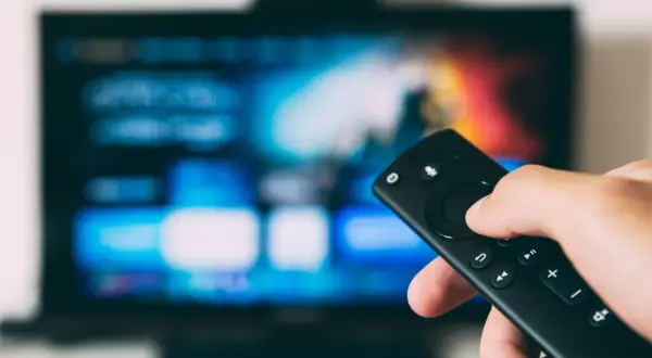 Dłoń trzymająca pilot do telewizora LG, ekran świeci w tle. Dowiedz się, jak wyłączyć lektora w telewizorze LG.