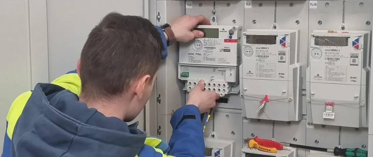 Technik w kamizelce wykonuje wymianę licznika Tauron. Widać nowe liczniki i narzędzia.