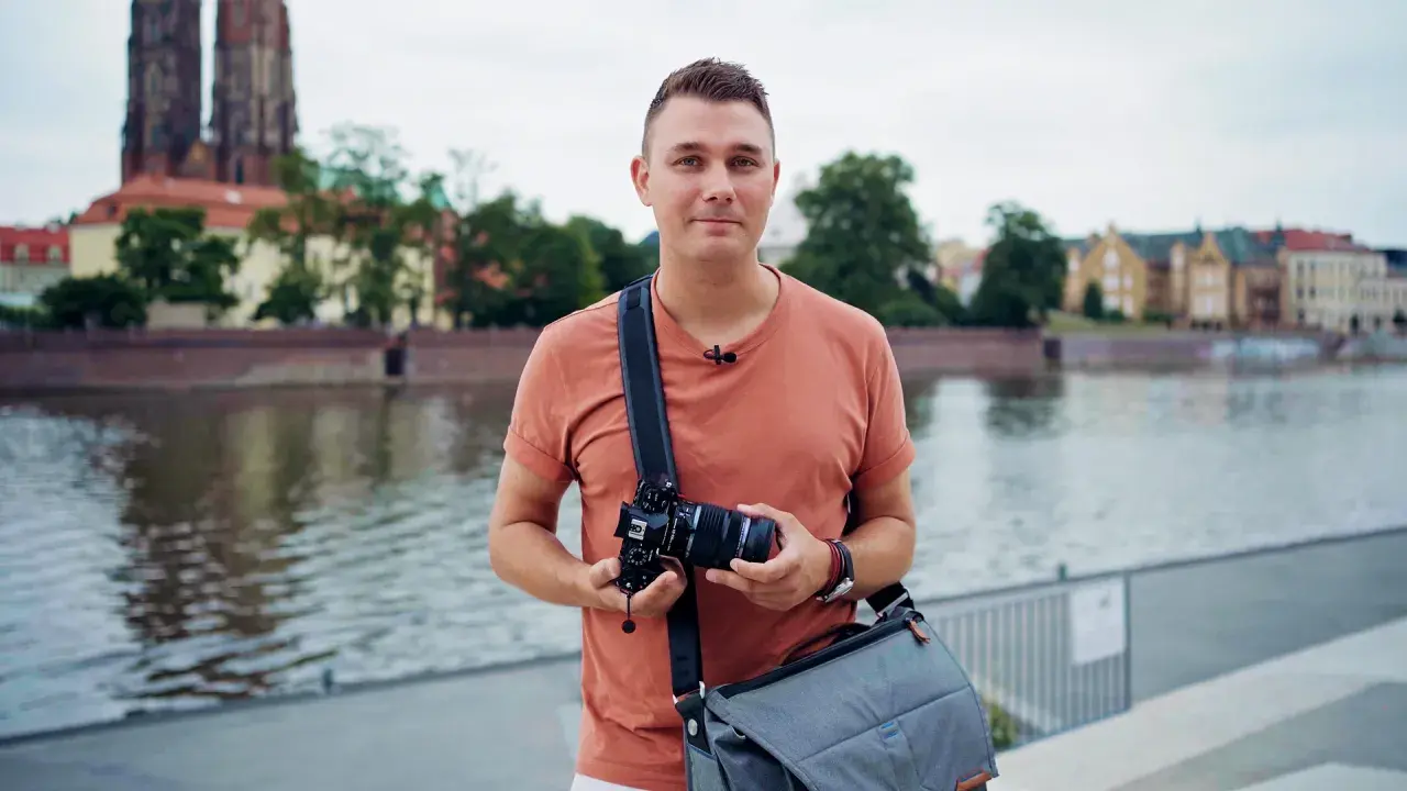 Mężczyzna w pomarańczowej koszulce trzyma aparat fotograficzny na tle rzeki i zabytkowych budynków.