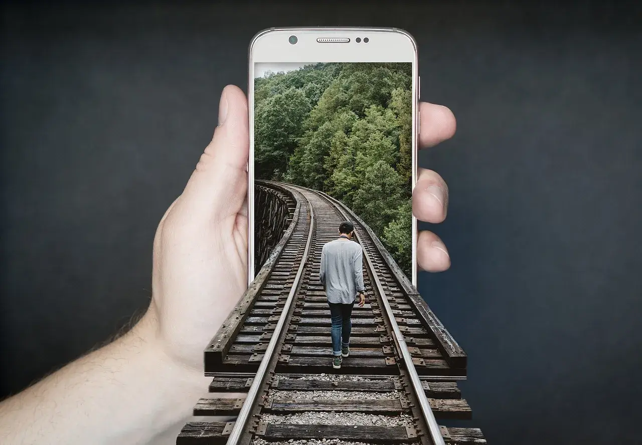 Dłoń trzyma telefon z ekranem **amoled**, na którym widać mężczyznę idącego po torach kolejowych przez las.
