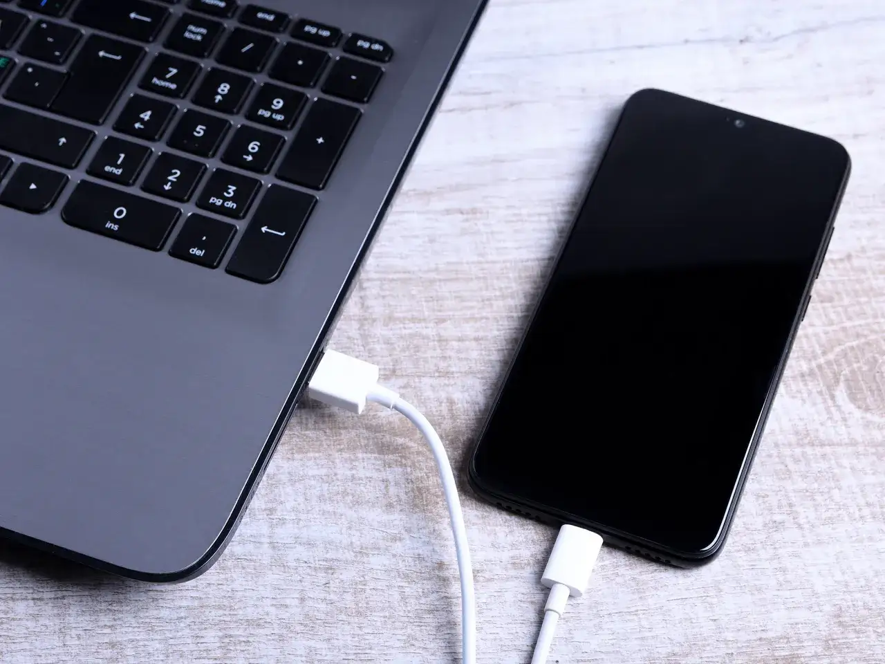 Jak połączyć telefon z komputerem przez USB? Biały kabel łączy czarny smartfon z szarym laptopem.