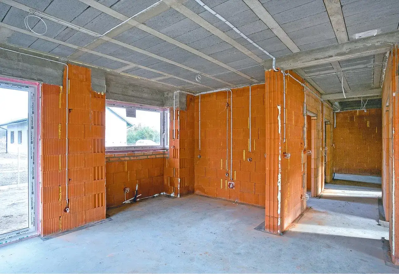 Wnętrze domu w budowie z cegły ceramicznej, betonowym sufitem i oknami. Widoczne instalacje elektryczne, przygotowane do dalszych prac. WLZ w trakcie montażu.