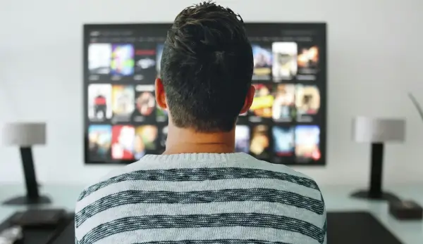 Mężczyzna w pasiastym swetrze ogląda na telewizorze menu aplikacji, gdzie widać okładki filmów i seriali.