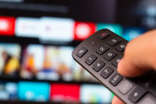 Dłoń trzyma pilot, a na ekranie telewizora widać kolorowe kafelki z filmami. Zastanawiasz się, iptv co to? To przyszłość telewizji!