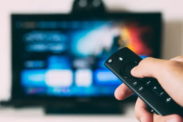 Dłoń trzymająca pilot do telewizora LG, ekran świeci w tle. Dowiedz się, jak wyłączyć lektora w telewizorze LG.