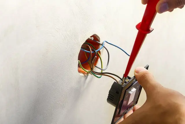 Instalator podłącza włącznik światła do puszki, używając śrubokręta. Widać przewody elektryczne wychodzące ze ściany.